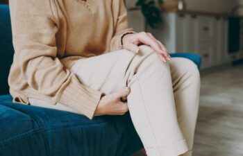 Cropped close up photo of pensioner woman wearing casual clothes sit on blue sofa hold knee suffer from leg knee pain inflammation stay home flat spend free spare time in living room indoor grey wall