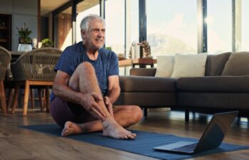 Fitness, laptop and senior man with ankle pain, injury or accident with virtual exercise or training. Computer, health and elderly male person with muscle sprain for online workout in living room.